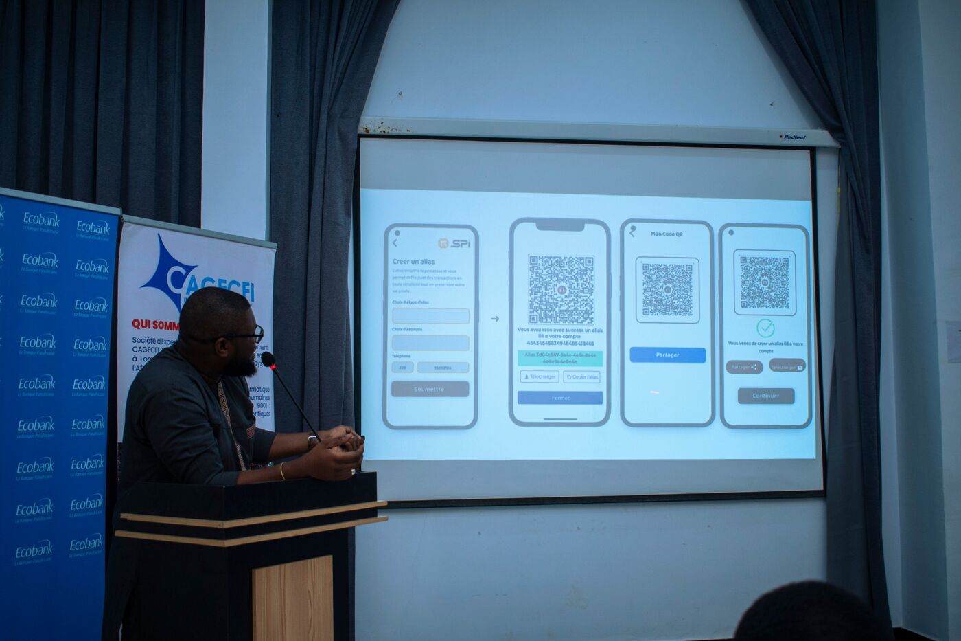 Eklin at the lectern looking at a projected slide showing the PI-SPI alias creation and QR code screens, with Ecobank and CAGECFI banners