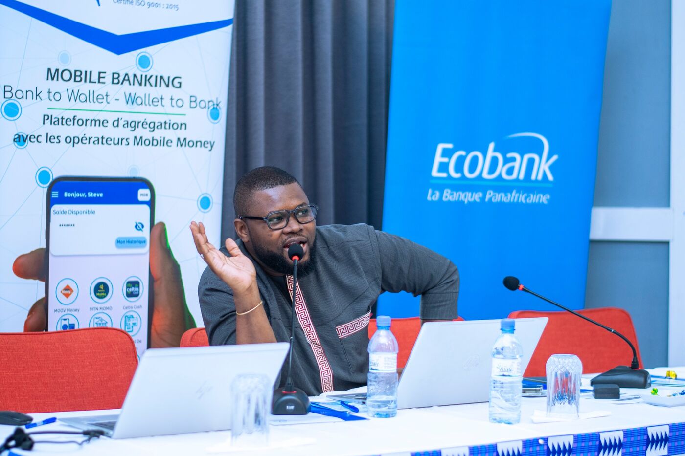 Eklin speaking at a table with a microphone, next to Mobile Banking and Ecobank banners