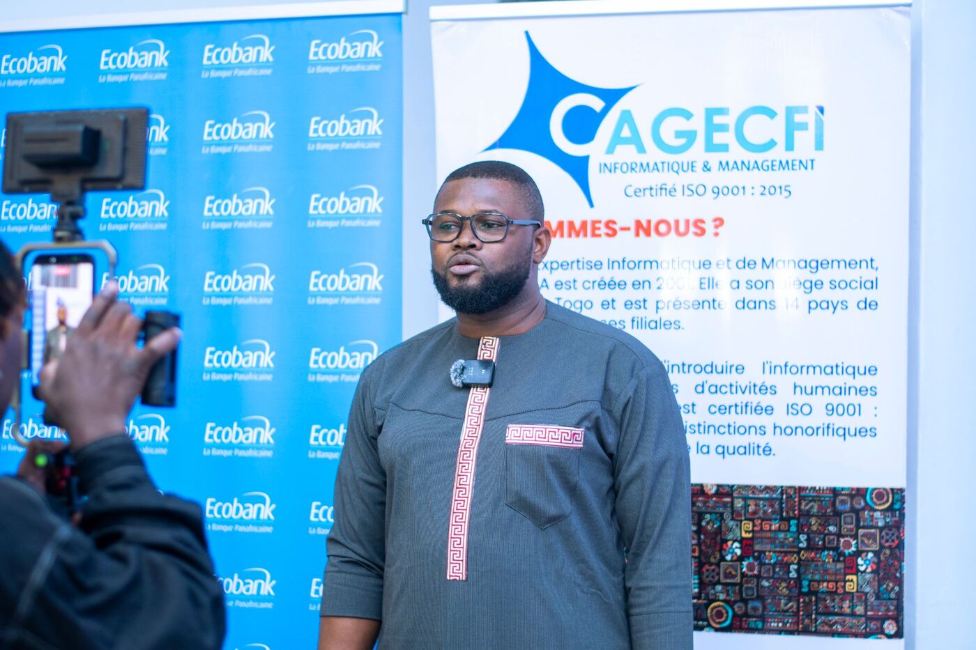 Eklin being filmed during an interview, in front of Ecobank and CAGECFI banners